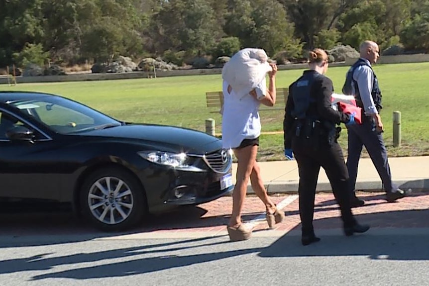 A woman holding a pillow across her face walks with police across a road near a park.