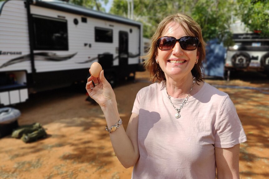 A woman holds up an egg in front of a caravan