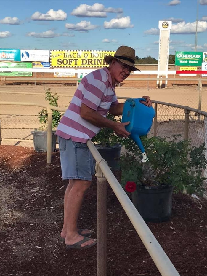 Man dressed casually watering his pot plants with dry race course behind him