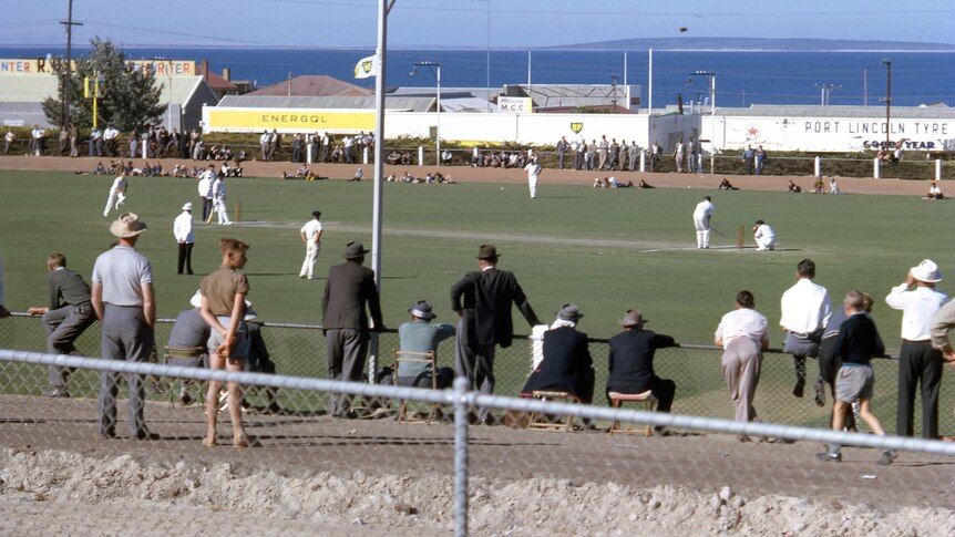 A game of cricket is played on an oval, as seen from the stands, beneath a sunny sky.
