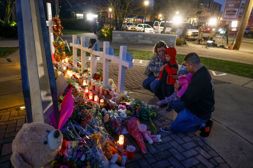 A man, a woman and two small children kneeling at a shrine of flowers, candles and five crosses. 