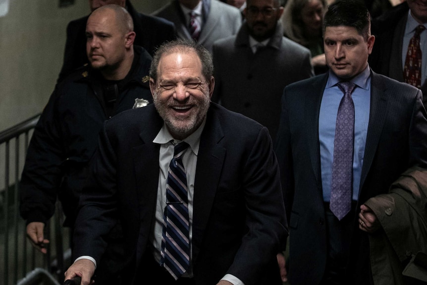 Harvey Weinstein walks through a hallway, smiling, while wearing a dark suit and tie.