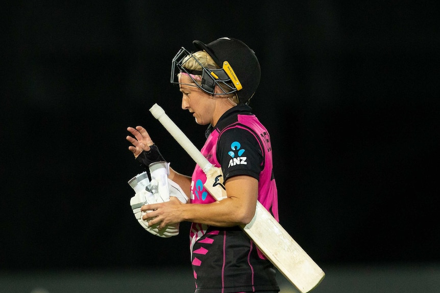 A woman walks off with her helmet angled on the top of her head and her bat under her arm