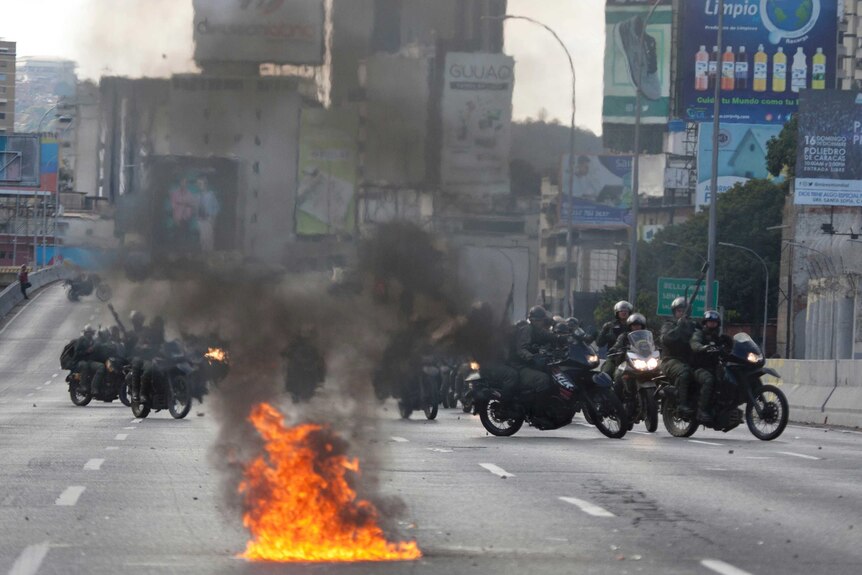 About a dozen police in black on motorbikes on a road with fire burning in the foreground.