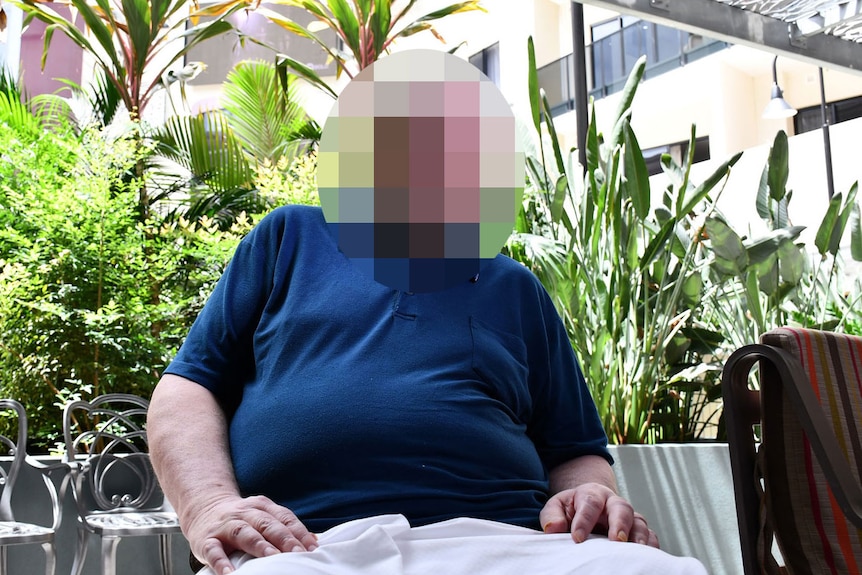 A man sits in a garden outside a city building, his face blurred to protect his identity.