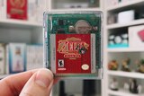 Close up a two fingers holding up a small games card with 'Zelda' written on it, and blurred shelving behind it.