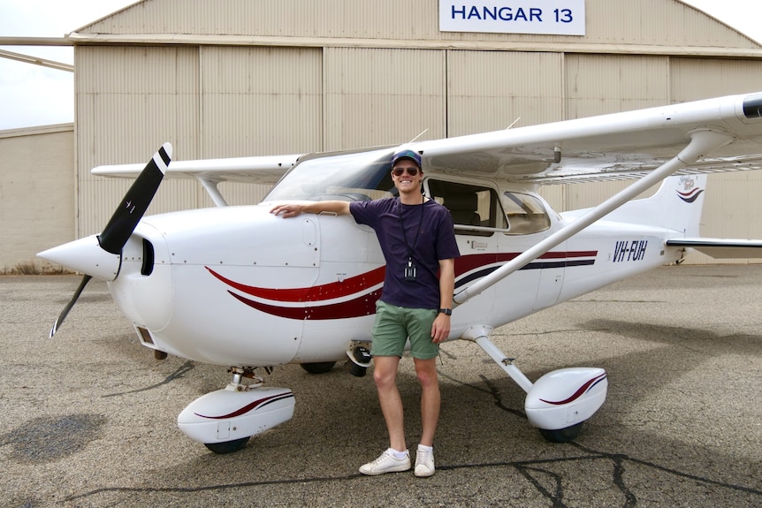 A young man smiles as he rest his arm on the body of a small plane.
