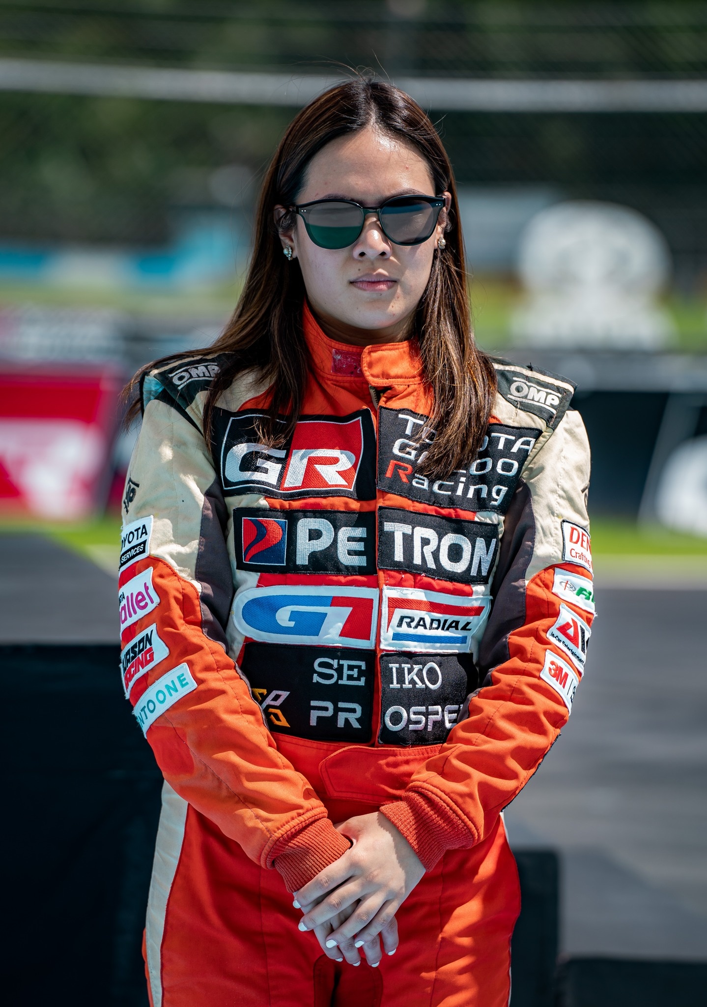 A woman in racing gear and sunglasses looks seriously at the camera.