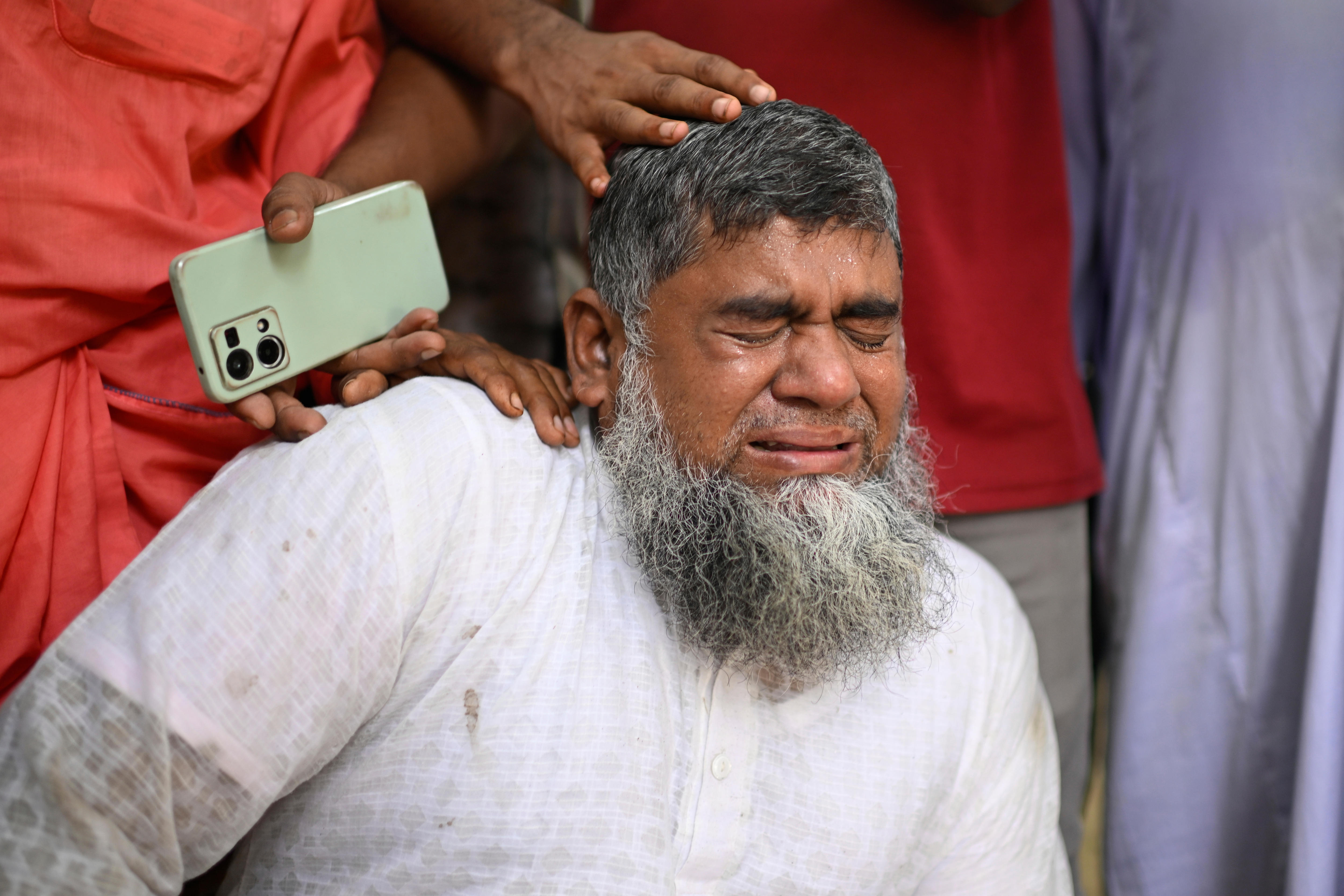 A Bangladeshi man with a greying beard wearing a white top crying while someone places a hand on his head