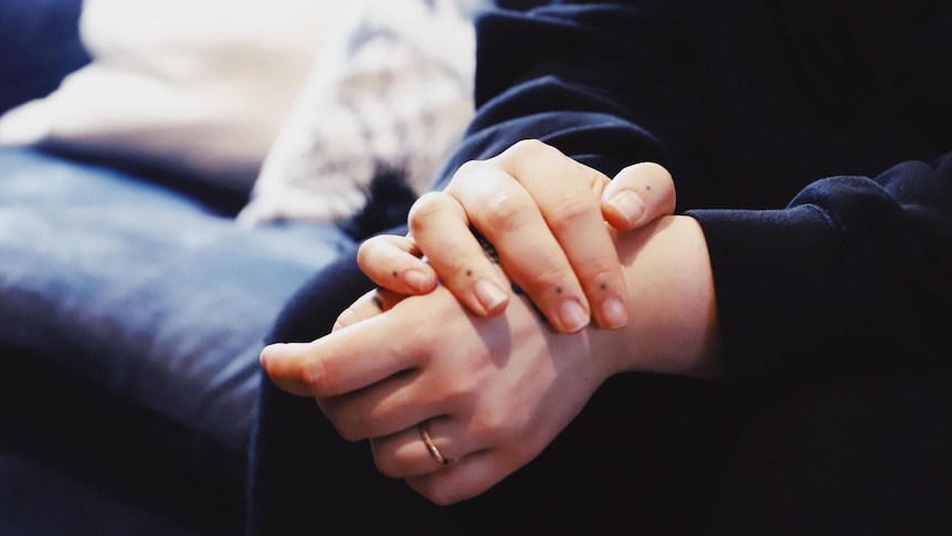 A close-up photo of a woman holding her hands together with a blue couch in the background.