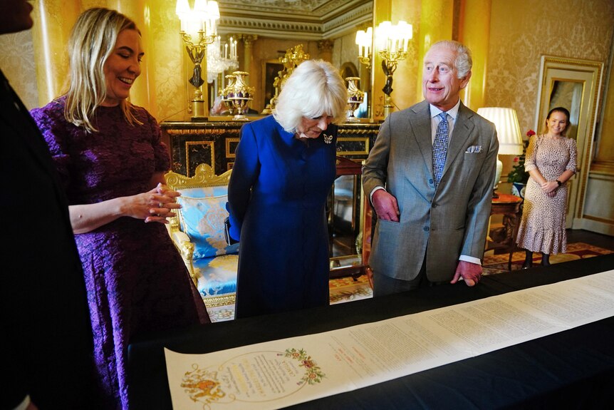 King Charles and his wife Camilla inspect a long roll of paper in an ornate room