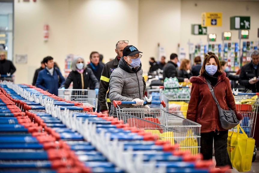 A line of people wearing masks and winter clothes push shopping trolleys in a crowded supermarket.