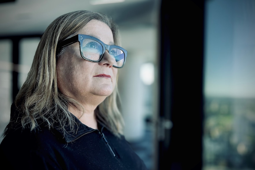 A middle aged woman with a blonde bob and thick rimmed glasses stands near a window looking into the distance.