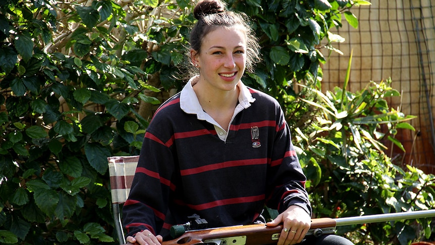 Am 18 year old girl sits on a camp chair in a garden with a rifle across her lap.