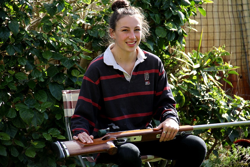 Am 18 year old girl sits on a camp chair in a garden with a rifle across her lap.