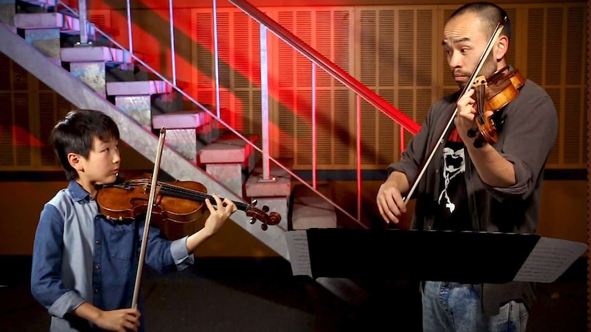 A boy playing a violin and a man playing a viola look at each other while playing in front of a beautifully lit staircase.