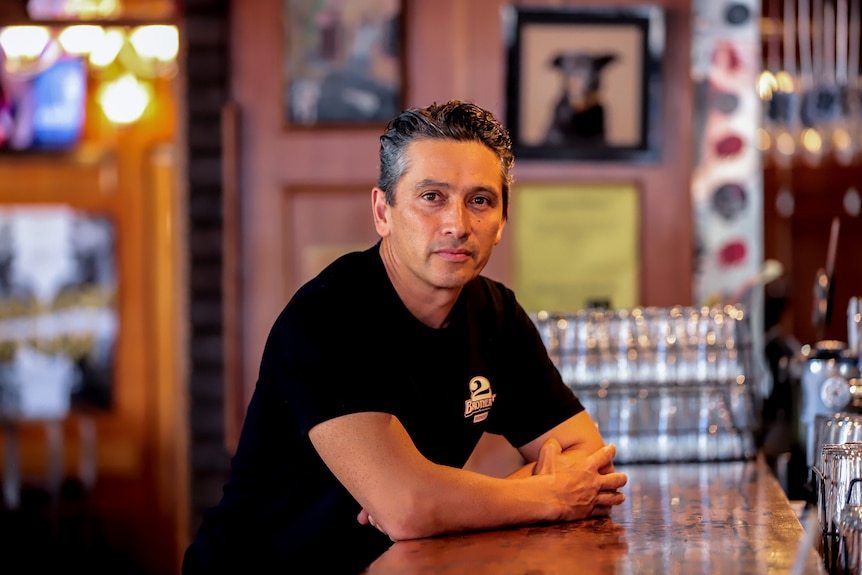 Man wearing black t-shirt leans over bar inside timber-walled pub