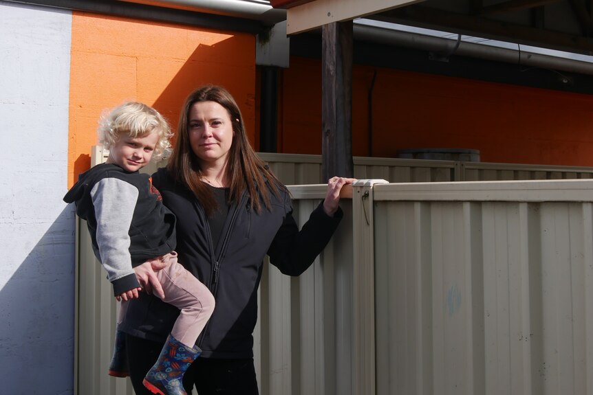 A serious woman holds a child next to a fence in front of an orange wall.