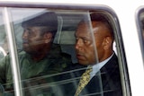 Two men sit in a car with the windows up, one wearing a suit the other a military uniform.