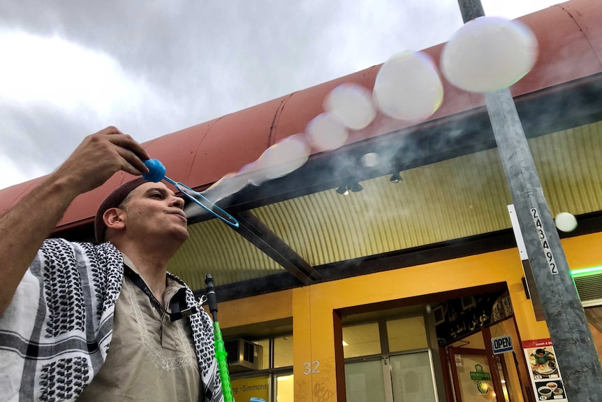 Wal Eade holds a bubble wand as he blows bubbles with smoke inside them. He is seen from below, with a grey sky above.