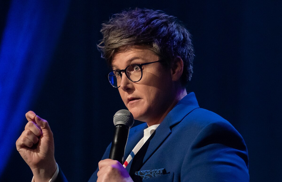 Comedian with short hair and glasses holding mic and gesturing with hand.