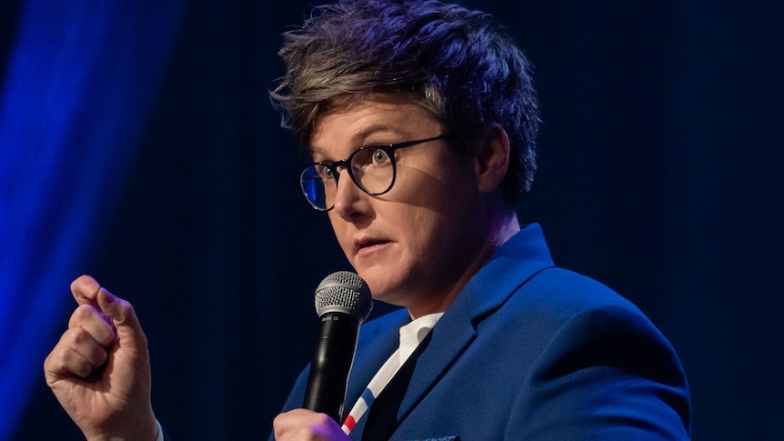 Comedian with short hair and glasses holding mic and gesturing with hand.