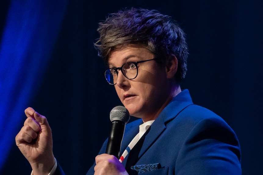Comedian with short hair and glasses holding mic and gesturing with hand.
