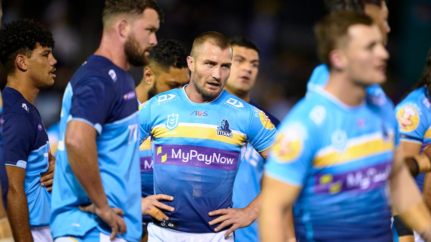 A Gold Coast NRL player stands with hands on hips amongst his teammates before a game.