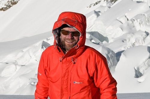 Huw Kingston stands in knee-deep snow, wearing an orange jacket and holding a camera.
