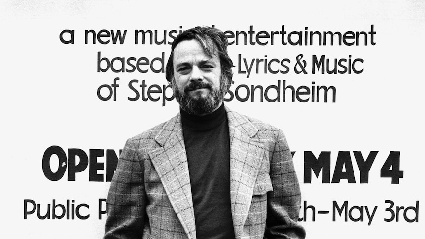 A black and white photo of Stephen Sondheim posing in front of a poster for one of his shows.