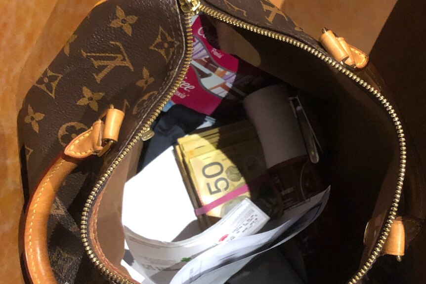 A brown patterened overnight bag with a stack of $50 bills in a rubber band spilling out.