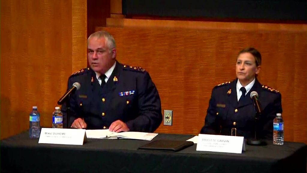 A police officer speaks at a microphone while another officer sits beside him.