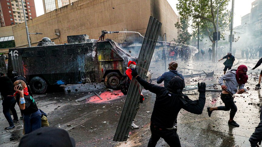 A chaotic street scene shows a paint-splattered military vehicle spraying water to ward off hooded protesters.