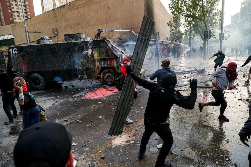 A chaotic street scene shows a paint-splattered military vehicle spraying water to ward off hooded protesters.