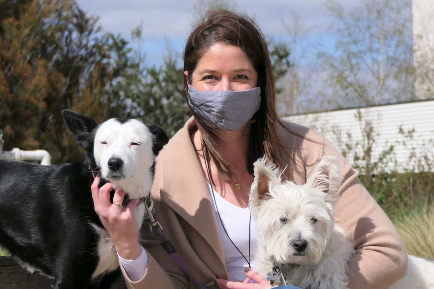 A woman looks into the camera wearing a mask. She holds a small white dog and a black and white larger dog.