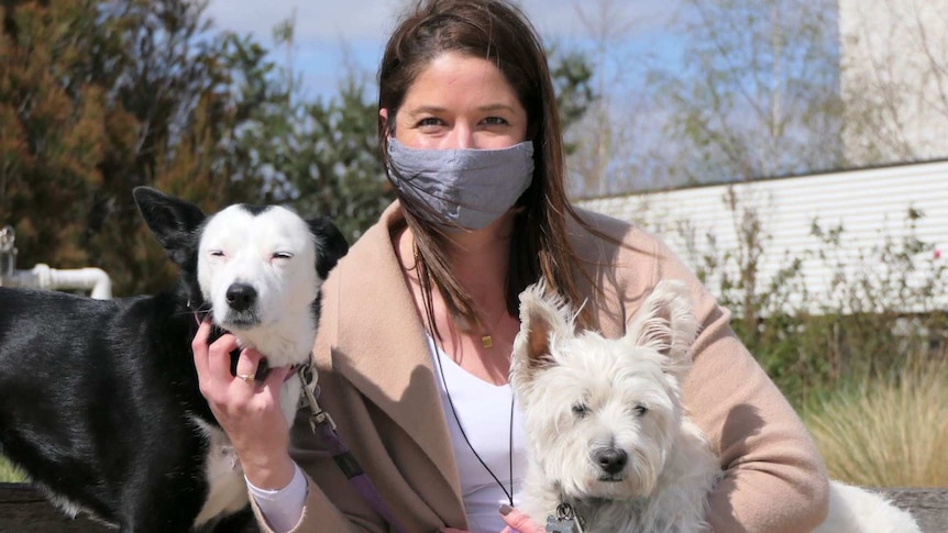 A woman looks into the camera wearing a mask. She holds a small white dog and a black and white larger dog.