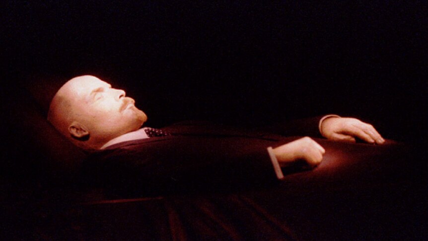 The dead body of Vladimir Lenin, a bald man with pointy beard, is propped up, his eyes closed and hands resting