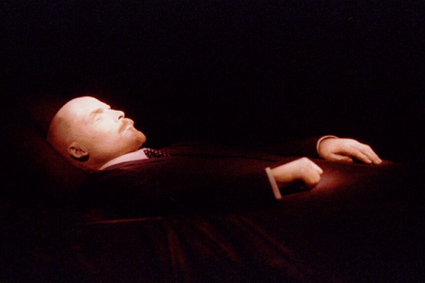 The dead body of Vladimir Lenin, a bald man with pointy beard, is propped up, his eyes closed and hands resting