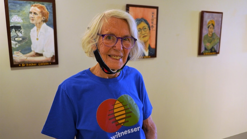 A woman wearing a blue t-shirt and with a face mask on her chin stands in front of three paintings on a wall