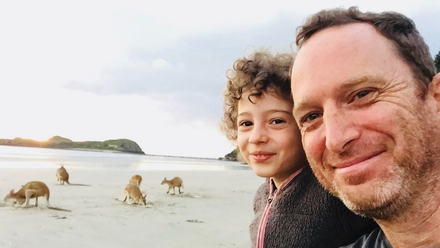 A man and young girl in a selfie with kangaroos on a white beach, with blue ocean, behind them.