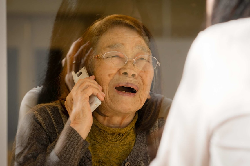 A woman with brown hair and glasses stares sadly at a woman behind a glass screen as she holds a telephone to her ear.