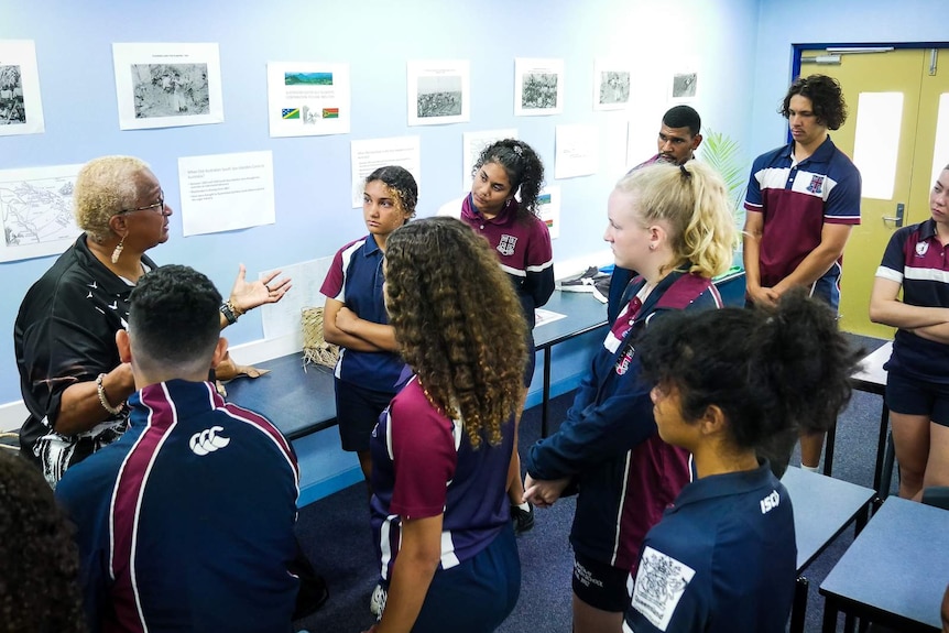 Woman speaking with a class of high school students with pictures and artefacts on the wall behind.