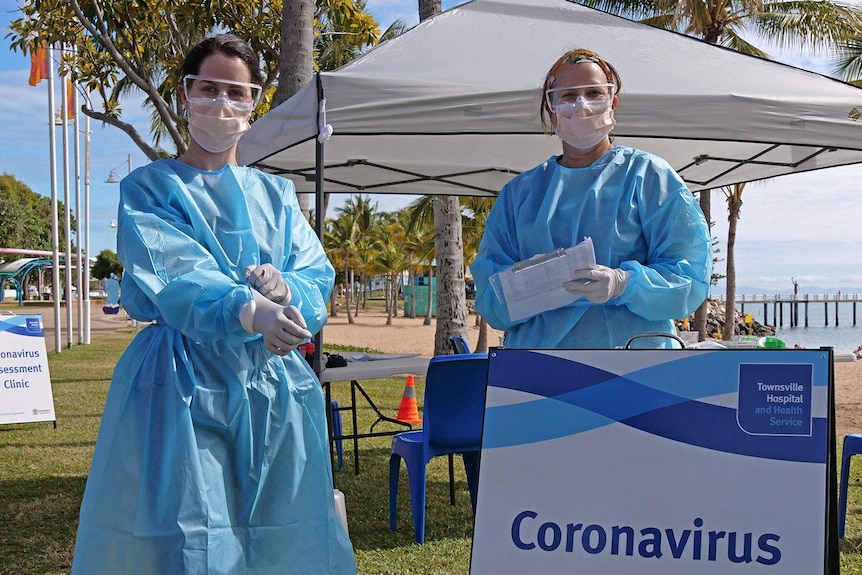 Two nurses wearing protective gear and masks stand in front of a tent on Townsville's foreshore, the beach is in the background