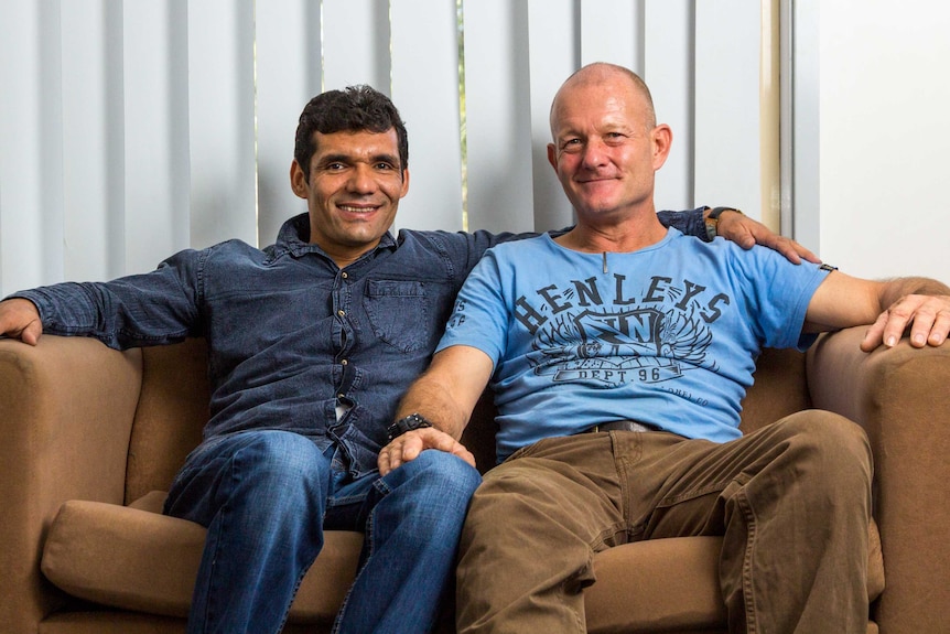 Jan, wearing a dark blue shirt and jeans, with Simon Quaglia, in a blue T-shirt and brown trousers, sitting on a couch