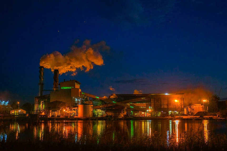 A landscape shot of a sugar mill lit up at night, with smoke rising from its chimneys.