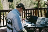 A person with denim jacket and cap seen from behind sitting outdoors at a table looking at phone with laptop open.