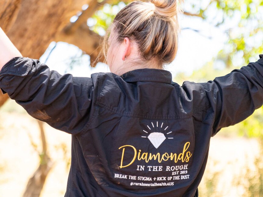 A woman stands with her back to the camera, wearing a jacket that reads: "Diamonds in the Rough. Est 2019. Break the stigma."