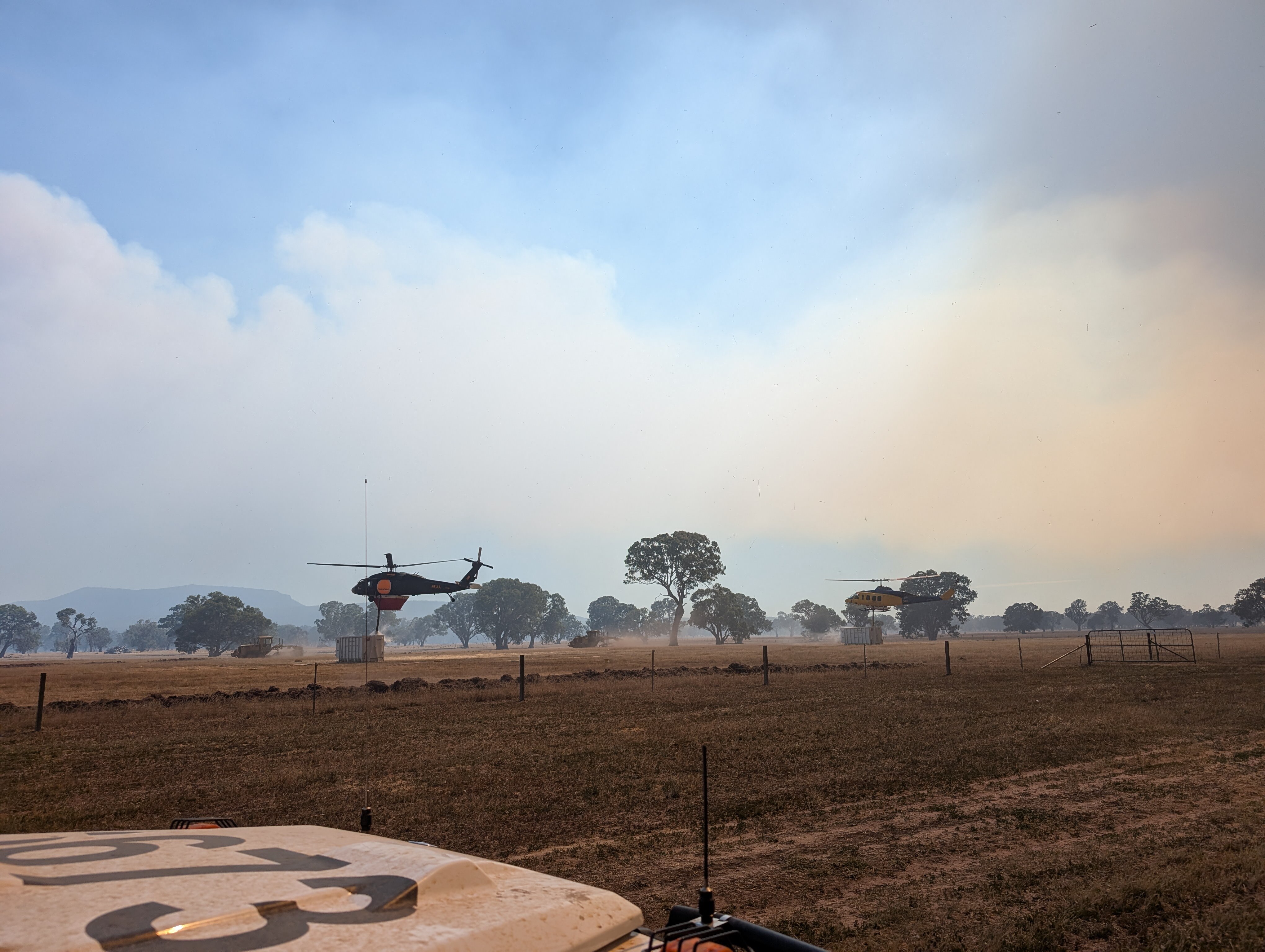 Two helicopter hover above water storage tanks in a dusty paddock with smoke in the background.
