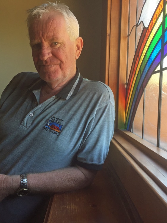 Phil Walcott at his home in Alice Springs, behind him is a rainbow themed stain glass window
