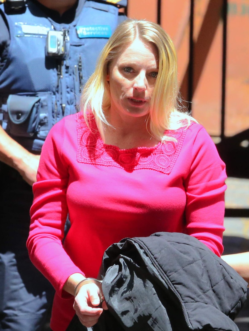 A handcuffed woman in a pink top carries a black jacket.